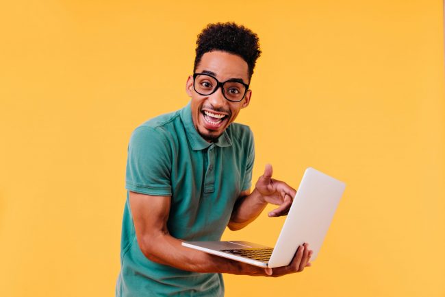 Blithesome student in green t-shirt posing with laptop. Indoor photo of amazed male freelancer isolated on yellow background.